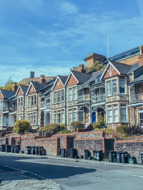 Exterior view of a row of multi-storey Victorian terraced houses with decorative white wrought iron balconies and large bay windows, situated on a slightly elevated brick terrace with steps leading up to each property. The houses have brick facades with contrasting white details and are set against a clear blue sky. In the foreground, there is a paved street with black waste bins lined along the pavement, which is adjacent to the residential buildings. Natural daylight illuminates the scene, highlighting the architectural features. This setting reflects a typical residential area suitable for house removals and home relocation services provided by Man with Van West Wickham, with the scene indicating potential activity involving furniture transport and loading processes.