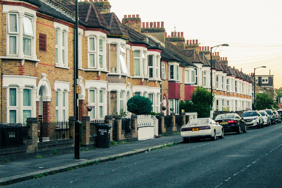 Photograph of a row of terraced houses in West Wickham, with brick facades and white-framed bay windows. The houses are situated along a paved sidewalk, separated by low brick and iron fences. Several landscaped front gardens contain small trees and shrubs. Parked cars line the street in front of the houses, including a silver vehicle parked on the pavement and others along the curb. The image is taken during daylight with soft natural lighting, capturing a quiet residential area suitable for house removals or relocation services. Man with Van West Wickham may use such areas for loading and unloading furniture during home relocation processes, with the street’s calm environment facilitating efficient furniture transport and packing activities inside the property.
