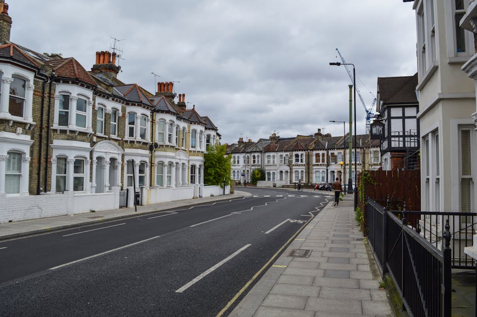 A quiet residential street in West Wickham featuring a row of Victorian-style terraced houses with white facades, bay windows, and red-tiled roofs. The pavement is lined with black metal fences and planters, with a few pedestrians walking along the sidewalk. In the background, there is a construction crane, indicating ongoing building work, and the sky is overcast with grey clouds. The scene captures the typical environment for home relocation and furniture transport services, with no vehicles or furnishings visible in the image, but the setting reflecting the area where professional removals like those provided by Man with Van West Wickham may operate for house moves or packing and moving tasks.