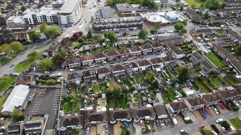 An aerial view of a residential neighbourhood in West Wickham with rows of terraced houses, each with small gardens and driveways, some featuring decking and patio furniture. The street is lined with parked cars and bordered by green lawns, trees, and shrubbery. Adjacent to the neighbourhood, there is a commercial building with a parking lot, and a larger multi-storey complex with flat roofs and white exterior walls. Visible in the foreground are several houses being prepared for home relocation, with furniture and boxes placed outside, some wrapped in plastic or fabric coverings, and a man with van vehicle nearby. The image illustrates a typical scene of furniture transport and packing in the context of house removals, emphasizing the logistics involved in loading and moving belongings within a residential area, supported by Man with Van West Wickham's removal services, as referenced in the web page about Bromley Council permits for West Wickham removals.