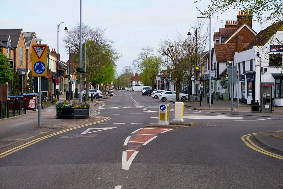 A quiet urban street scene in West Wickham with a roadblock in the foreground featuring two yellow and white traffic bollards and a small central island with a blue circular sign directing vehicles to keep left for a roundabout. To the left, a street sign indicates a give way priority. There are several parked cars along both sides of the street and a row of mixed residential and commercial buildings, including shops and houses, with brick and white facades. Trees with budding green leaves line the pavement, and lampposts are evenly spaced along the sidewalk. The sky is overcast, and the street appears to be suitable for a home relocation or moving logistics process managed by a professional removals company such as Man with Van West Wickham, who may be coordinating vehicle access and parking permits for furniture transport and packing and moving activities during a house removal in the area.