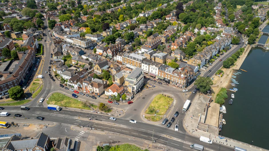 An aerial view of a residential neighbourhood in West Wickham with rows of terraced houses, each with small gardens and driveways, some featuring decking and patio furniture. The street is lined with parked cars and bordered by green lawns, trees, and shrubbery. Adjacent to the neighbourhood, there is a commercial building with a parking lot, and a larger multi-storey complex with flat roofs and white exterior walls. Visible in the foreground are several houses being prepared for home relocation, with furniture and boxes placed outside, some wrapped in plastic or fabric coverings, and a man with van vehicle nearby. The image illustrates a typical scene of furniture transport and packing in the context of house removals, emphasizing the logistics involved in loading and moving belongings within a residential area, supported by Man with Van West Wickham's removal services, as referenced in the web page about Bromley Council permits for West Wickham removals.
