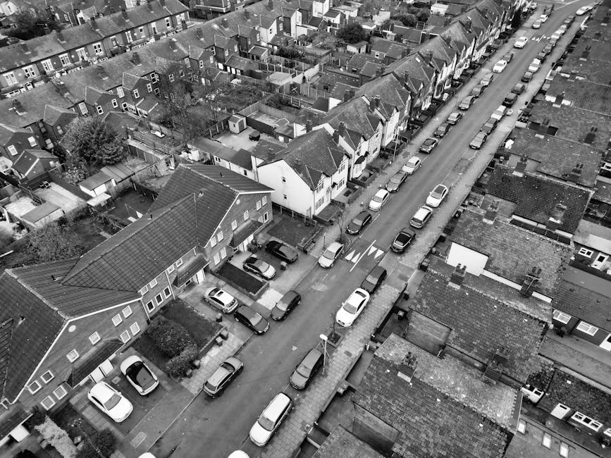 An aerial black-and-white photograph showing a residential street with parked cars lining both sides of the road, adjacent to rows of terraced houses with pitched roofs and small backyards. The street features a designated loading area with a collection of cardboard boxes, pieces of furniture covered with protective blankets, plastic wrap, and packing materials, positioned near the entrance of a house. A man with a van from Man with Van West Wickham, visible on the right side of the image, is engaged in a home relocation process, loading or unloading furniture and boxes onto the vehicle using trolleys and straps. The scene illustrates the logistics involved in furniture transport and packing for house removals within a typical suburban neighborhood, emphasizing careful handling, loading procedures, and the use of appropriate moving equipment to ensure safe and efficient home relocation services.