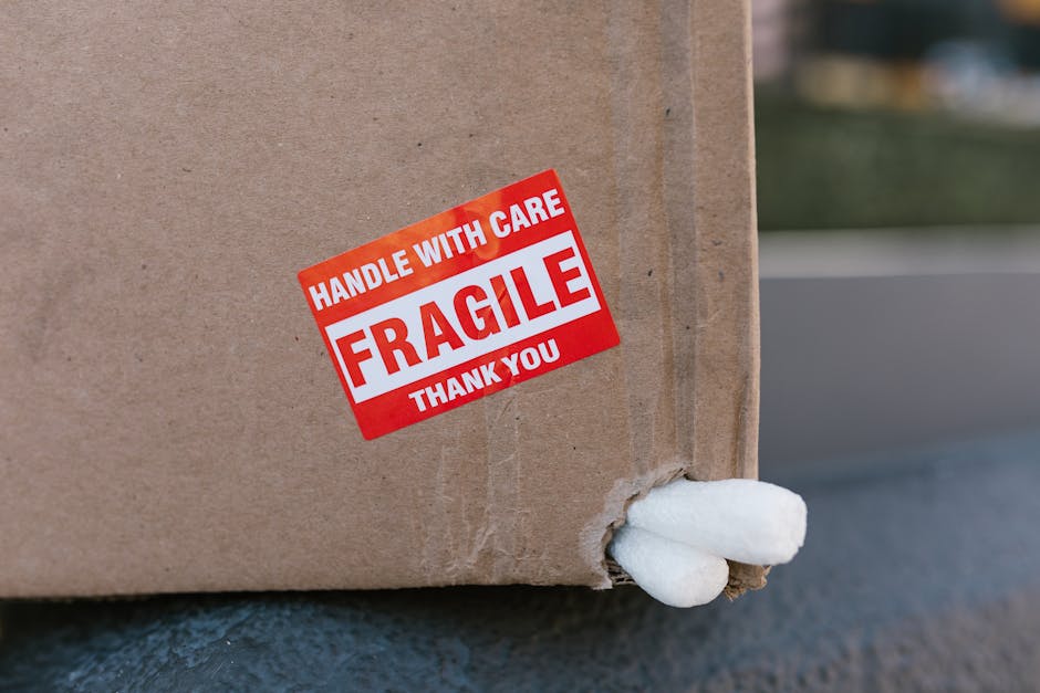 Close-up image of a cardboard box with a red and white 'Fragile' label that reads 'Handle with Care, Fragile, Thank You.' The box is positioned outside on a driveway or pavement, partially resting on a black surface with a small section of torn cardboard revealing packing material inside. The background shows a blurred outdoor environment with some greenery and distant buildings. The box appears to be part of a house removal or moving process managed by Man with Van West Wickham, with the label indicating that it contains delicate or valuable items requiring careful handling during the furniture transport or home relocation.