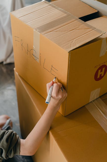 A close-up of a person's hand using a black marker to label a cardboard moving box with the word 'FRAGILE' written in capital letters. The box is sealed with brown packing tape and is placed on top of other packed boxes. The setting appears to be inside a home or a moving storage area, with natural light illuminating the scene. The background includes additional cardboard boxes, some stacked, and part of a padded surface, indicating preparations for home relocation. This image reflects the packing process involved in furniture transport and house removals, consistent with the services offered by Man with Van West Wickham, especially in relation to packing fragile items during a move.