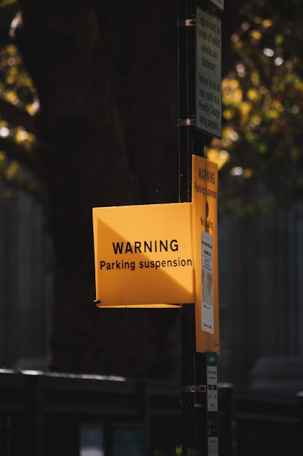 A close-up photograph of a yellow warning sign attached to a metal pole outdoors, indicating a parking suspension. The sign reads 'WARNING Parking suspension' in black text. Behind the sign, there are additional smaller notices and a partially visible green parking regulation sign. The background features blurred trees with leaves that have a warm, golden hue and a dark fence or wall, suggesting the scene is set in a residential or urban neighbourhood. The lighting indicates late afternoon or early evening. The image visually relates to the context of parking restrictions, which are relevant to house removals and transport logistics, as shown on the page about skipping parking suspensions for home relocations with Man with Van West Wickham.