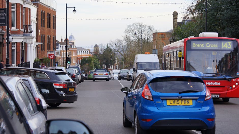 A busy urban street scene during daytime with multiple parked cars along the roadside, including a black sedan, a grey hatchback, and a white van, all aligned along the curb. In the foreground, a blue hatchback with a EU registration plate is parked closely to the pavement, while a red Metroline bus displaying the route number 143 towards Brent Cross is moving along the street. The background features a mix of brick and white buildings with entrance steps, street lamps, and leafless trees, suggesting a late autumn or early winter setting. The street is well-lit with natural daylight under an overcast sky, and the scene captures the typical environment for house removals or furniture transport operations, with visible space for loading and unloading. Occasionally, occasional pedestrians can be seen walking along the sidewalks, emphasizing the urban context that Man with Van West Wickham operates within for residential and commercial relocation services.