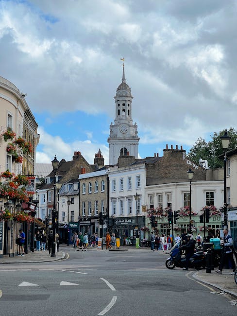 A street scene in West Wickham showing several residential and commercial buildings, some with flower baskets hanging outside. In the background, there is a tall clock tower with a weather vane on top, part of a historic town hall or church, rising against a partly cloudy sky with patches of blue. The foreground features people walking on the pavement and crossing the road, with some standing near shops, and vehicles including a motorcycle and a few cars parked or moving along the street. The area appears busy with activity typical of a community hub, during daylight. This setting can be associated with a location where house removals and furniture transport services might be carried out, emphasizing the context of urban logistics and moving operations by companies like Man with Van West Wickham.