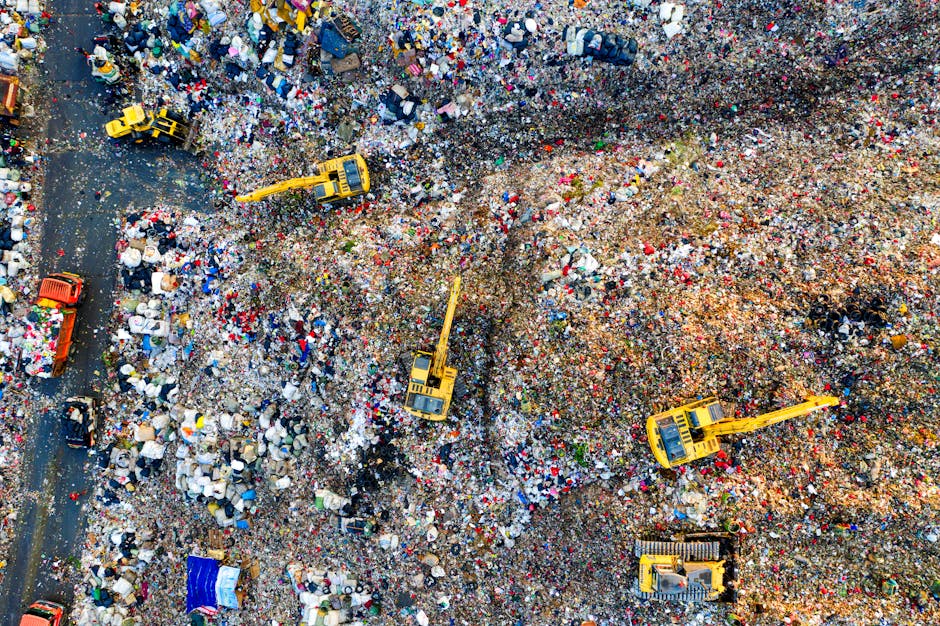 An aerial view of a large landfill site filled with various types of waste, including plastic, cardboard, textiles, and household rubbish. Several yellow excavators are actively working on sorting and moving the debris, with some using buckets attached to their arms, while others are positioned on different sections of the site. The waste is scattered across the uneven terrain, creating a mix of colors and textures, with piles of discarded materials spilling onto a nearby paved road. The scene is well-lit with natural daylight, highlighting the scale of the landfill and the machinery involved in waste management operations. This image illustrates the environmental impact of waste disposal and landfill handling, related to waste disposal rules and proper home disposal practices addressed in the context of house removals and relocation services by Man with Van West Wickham.