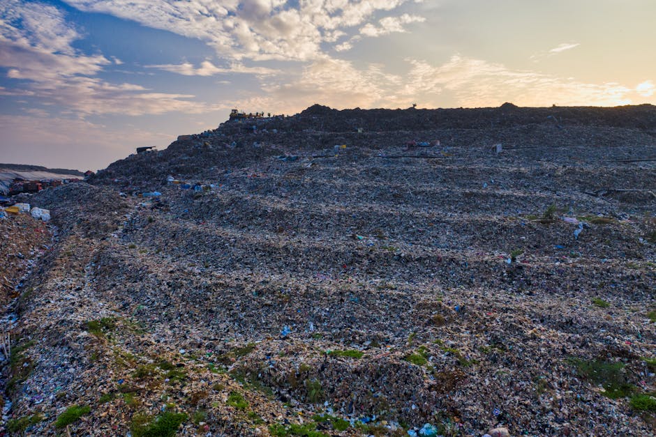 Photograph of a large landfill site at sunset, showing a vast area covered with layers of mixed waste materials, including plastic, paper, and debris. The landscape features terraced slopes created by accumulated trash, with scattered isolated patches of green vegetation. The sky above is partly cloudy with the sun casting a warm glow on the scene. In the background, a group of people can be seen standing on the top of the waste mound, observing the area. The landfill is situated next to a paved road, with some vehicles and equipment visible at the edges, indicating ongoing waste management or disposal activities. The image highlights the scale of waste accumulation in a landfill, emphasizing the importance of responsible waste disposal and recycling in home and furniture removal processes, as indicated in the context of waste disposal rules after a West Wickham move.