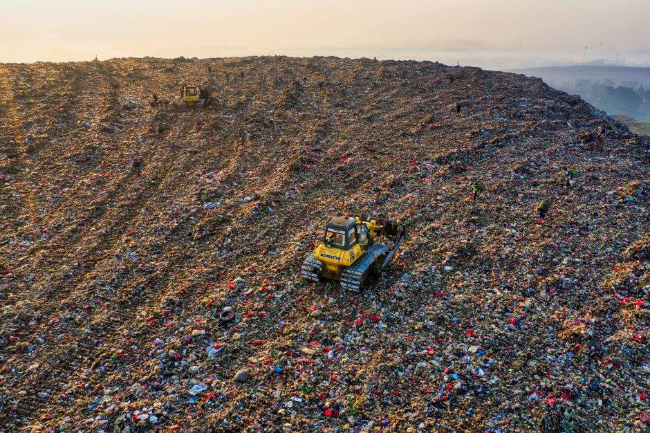 An aerial view of a large landfill site filled with various types of waste, including plastic, cardboard, textiles, and household rubbish. Several yellow excavators are actively working on sorting and moving the debris, with some using buckets attached to their arms, while others are positioned on different sections of the site. The waste is scattered across the uneven terrain, creating a mix of colors and textures, with piles of discarded materials spilling onto a nearby paved road. The scene is well-lit with natural daylight, highlighting the scale of the landfill and the machinery involved in waste management operations. This image illustrates the environmental impact of waste disposal and landfill handling, related to waste disposal rules and proper home disposal practices addressed in the context of house removals and relocation services by Man with Van West Wickham.