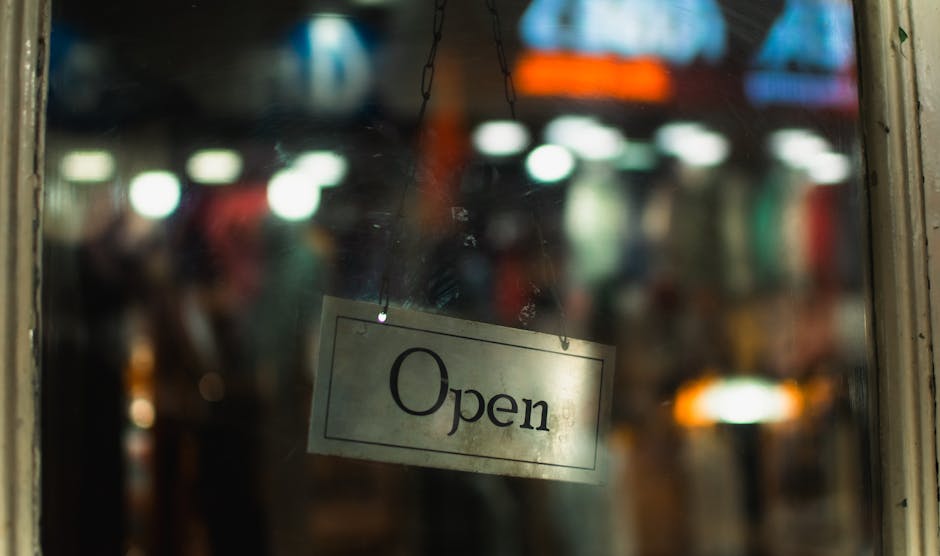 A close-up view of a glass door with a rectangular sign hanging from a chain, displaying the word 'Open' in black text on a white background. The door is part of a commercial property, likely a shop, with a blurred interior visible through the glass, showing various indistinct lights and objects. The door frame appears to be wood or metal, with some foliage or plant material along the right side. The lighting suggests daytime or well-lit premises, suitable for a retail location on a busy high street, and the scene relates to the environment where household or business relocations may occur, with [COMPANY_NAME] providing professional removals and transport services.