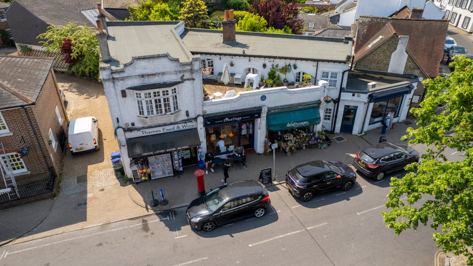 An aerial view of a small street with three retail shops in a white-painted building, featuring large front windows and awnings, situated in residential surroundings with trees and neighboring houses. The middle shop has a black awning, and the right-side shop has a green awning, both displaying menus or signs outside. Several parked cars are along the street, including a black hatchback, a dark SUV, and a white van parked on the pavement near the shops. The street pavement is visible in front of the shops, with people walking or standing nearby, possibly engaged in a home relocation or furniture transport activity, as part of a professional removals process involving loading or unloading equipment from nearby vehicles, supported by the context of [COMPANY_NAME] providing move and packing services in West Wickham.